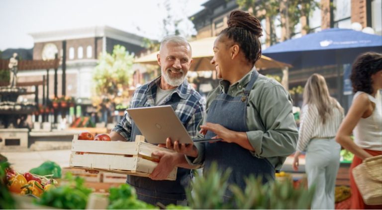 Two vendors chatting and holding a laptop at an outdoor farmers market.