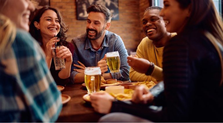 Friends laughing and toasting with beers at a restaurant table.