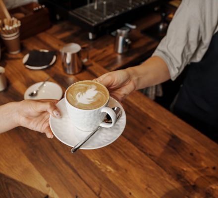 Barista handing a latte with foam art across a wooden counter.